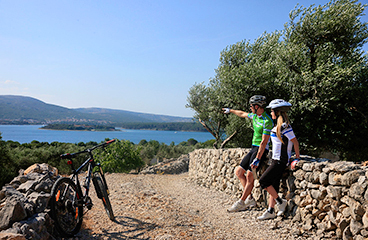 Un uomo e una donna si prendono una pausa, riposando contro un muro roccioso con le biciclette al lato. L'uomo indica il Mare Adriatico.