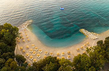 Aerial view of the pebbly Val Sunset Family beach, decked with sun loungers and umbrellas, surrounded by trees
