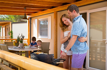 Ein Paar grillt auf der Terrasse eines Vista Mare Premium Campinghauses, während ihre Kinder im Hintergrund am Tisch spielen.