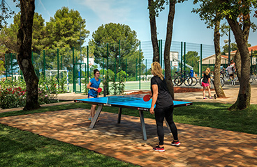 Dve ženski igrata namizni tenis v V Sportu v Lanterna Premium Camping Resortu z večnamenskim igriščem v ozadju