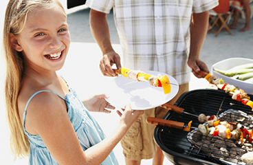 Een klein meisje lacht terwijl een man die aan het barbecueën is haar spiesjes serveert